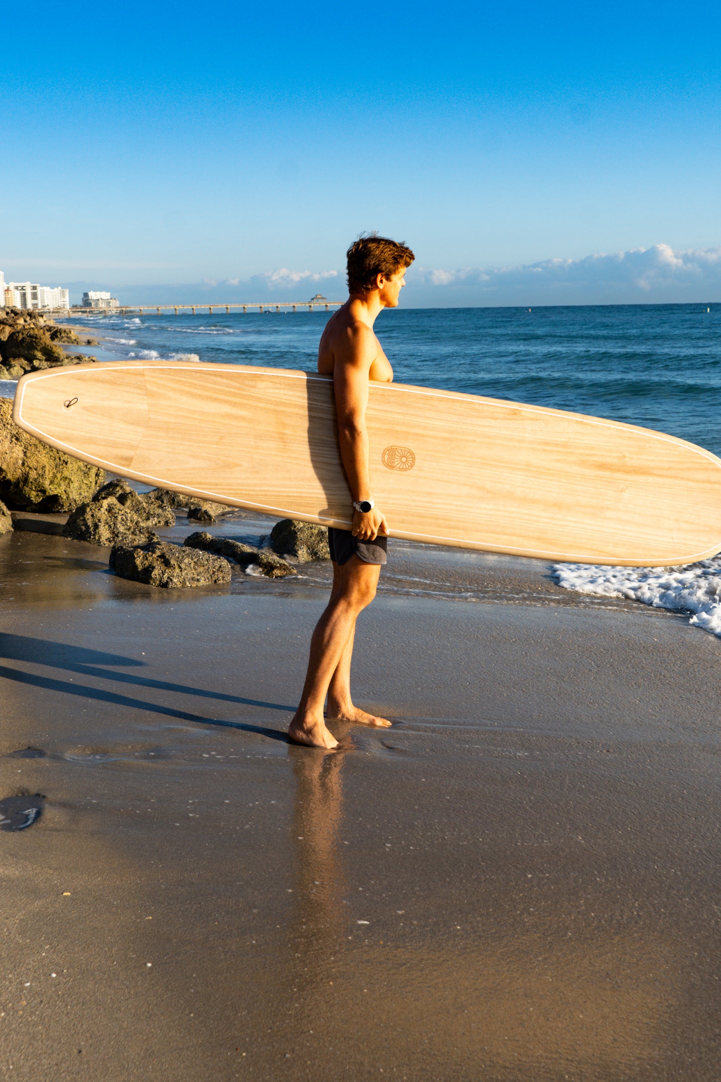 Man holding a wooden surfboard on a beach with ocean and sky in the background
