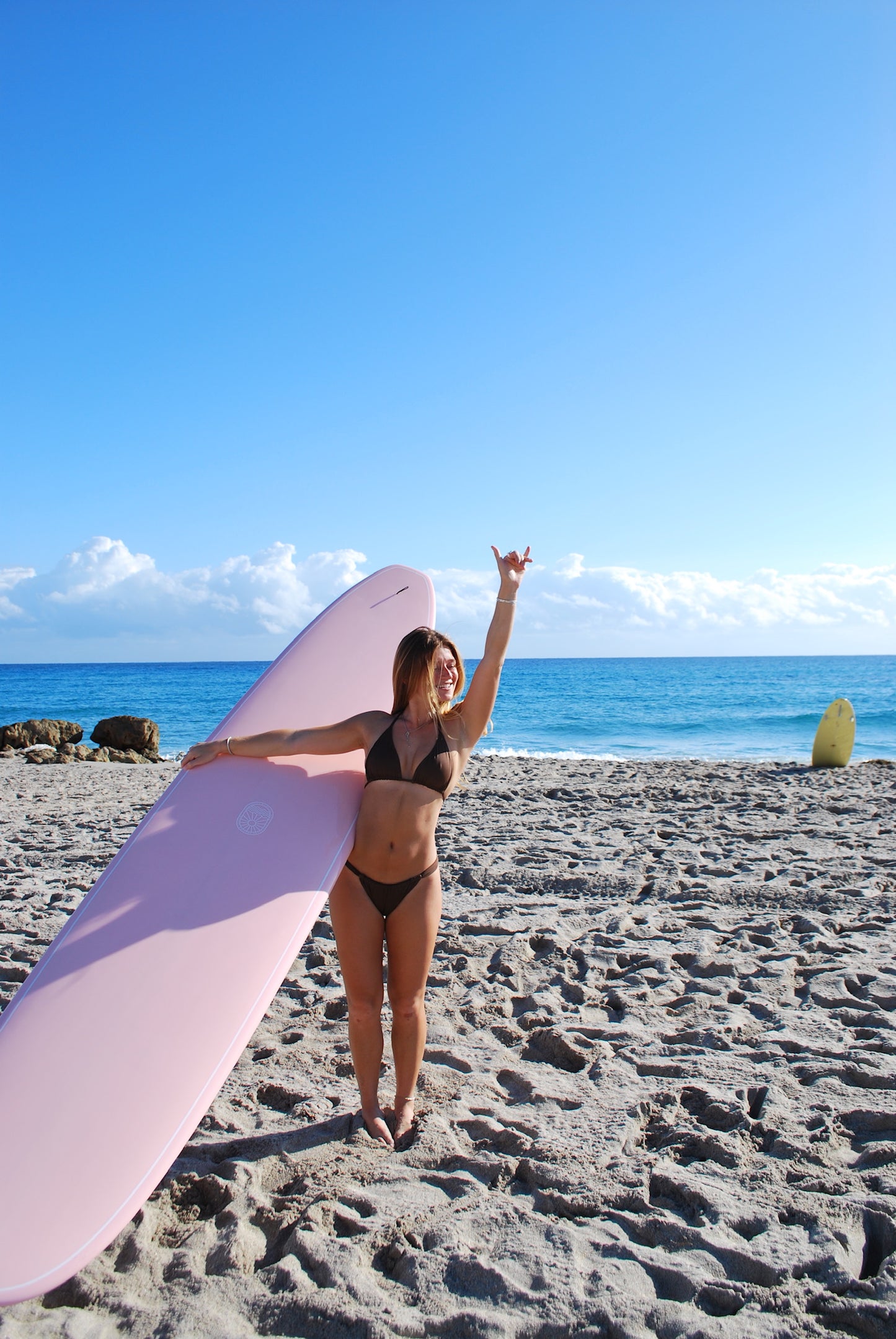 Woman holding a pink surfboard on a sandy beach with blue ocean and sky.