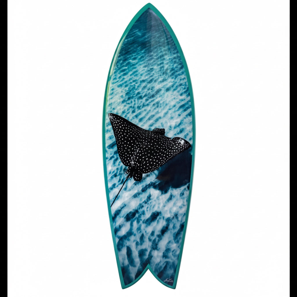 A surfboard featuring a blue ocean wave pattern with a black and white spotted stingray graphic in the center, set against a plain white background.